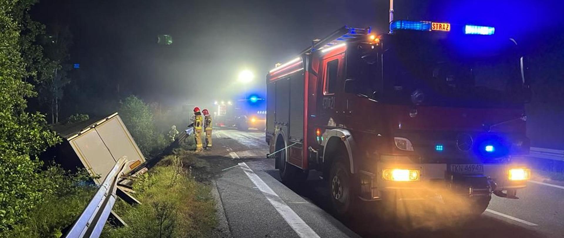 Wypadek ciężarowego DAF-a z osobową Hondą. Po prawej wóz strażacki po środku dwóch strażaków a po lewej w rowie tył naczepy samochodu ciężarowego który zjechał do rowu. Uszkodzona barierka i brzózki.