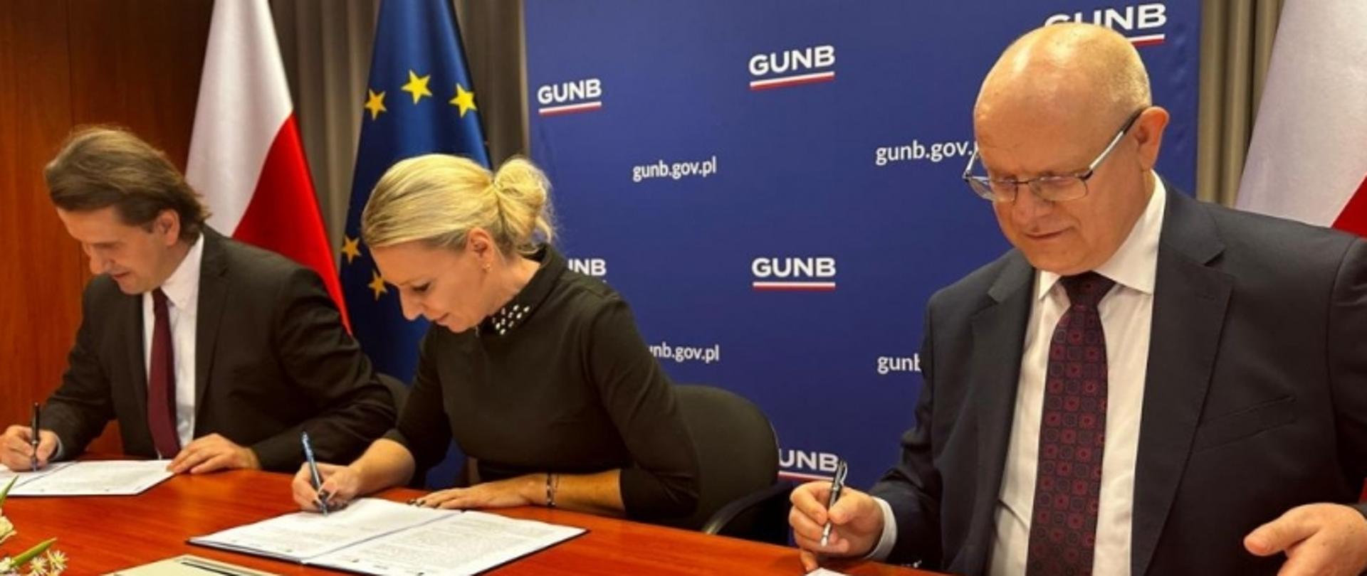 Trzy osoby podpisujące dokumenty na tle planszy „GUNB” i flag.