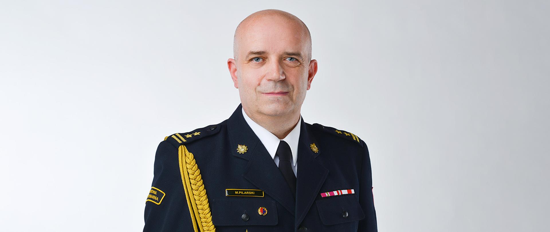 Zdjęcie przedstawia komendanta bryg. mgr inż. Marcina Pilarskiego na jasnym tle w mundurze galowym