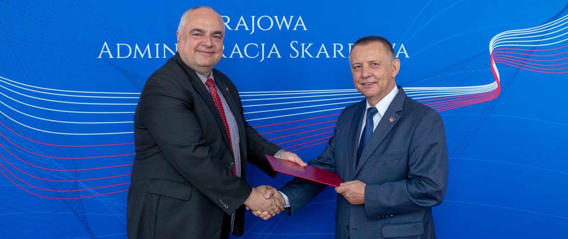 Uśmiechnięty minister Marian Banaś i wiceminister Tomasz Słaboszowski trzymają uściśniętą rękę na tle baneru Krajowej Administracji Skarbowej