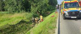 Po prawej stronie na ulicy stoi ZRM, po lewej stronie w rowie na trawie są strażacy dalej są drzewa , strażacy udzielają pomocy osobie poszkodowanej