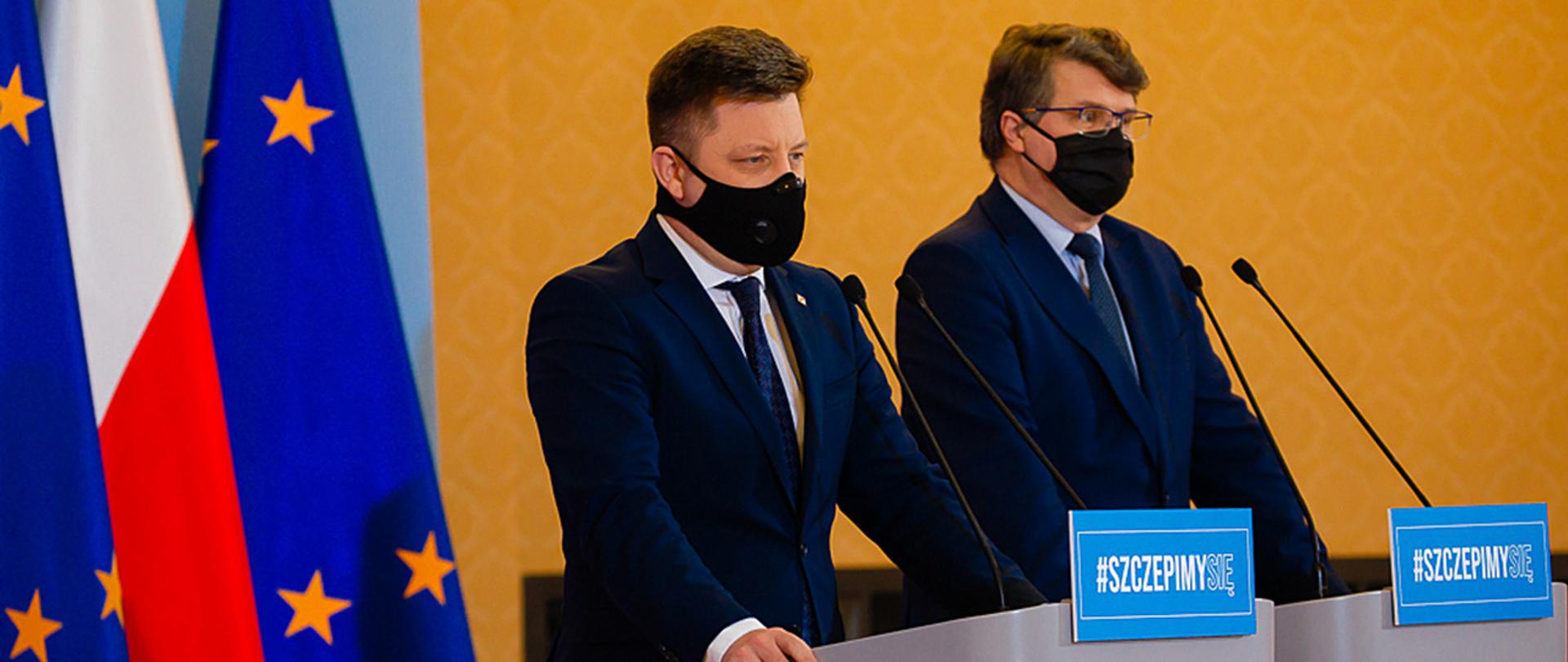 Na zdjęciu widać ministra Michała Dworczyk i wiceministra Macieja Wąsika stojących przy mównicach w trakcie konferencji prasowej. Minister Dworczyk zabiera głos. W tle widać flagi Polski i UE.