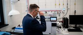 Minister Wieczorek stoi w pomieszczeniu z wyłożonymi białymi kafelkami ścianami, patrzy na monitor skomplikowanej maszyny stojącej na białym laboratoryjnym stole.