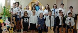 Grupa dzieci na zdjęciu grupowym, uczniowie klasy gitary trzymający gitary, na tle sceny auli PSM w Kłodzku, wraz z nauczycielami A. i W. Kozakowskimi