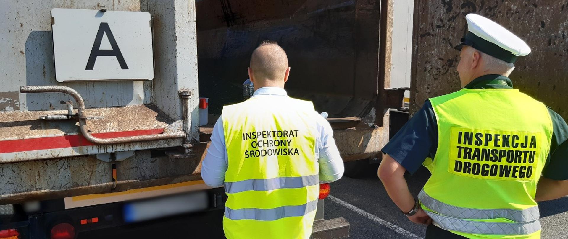 Od lewej: inspektor z Wojewódzkiego Inspektoratu Ochrony Środowiska i inspektor ze śląskiej Inspekcji Transportu Drogowego stoją za naczepą ciężarową, którą przewożone są odpady. Inspektorzy dokonują oględzin i oceny odpadów przewożonych w ładowni naczepy. 