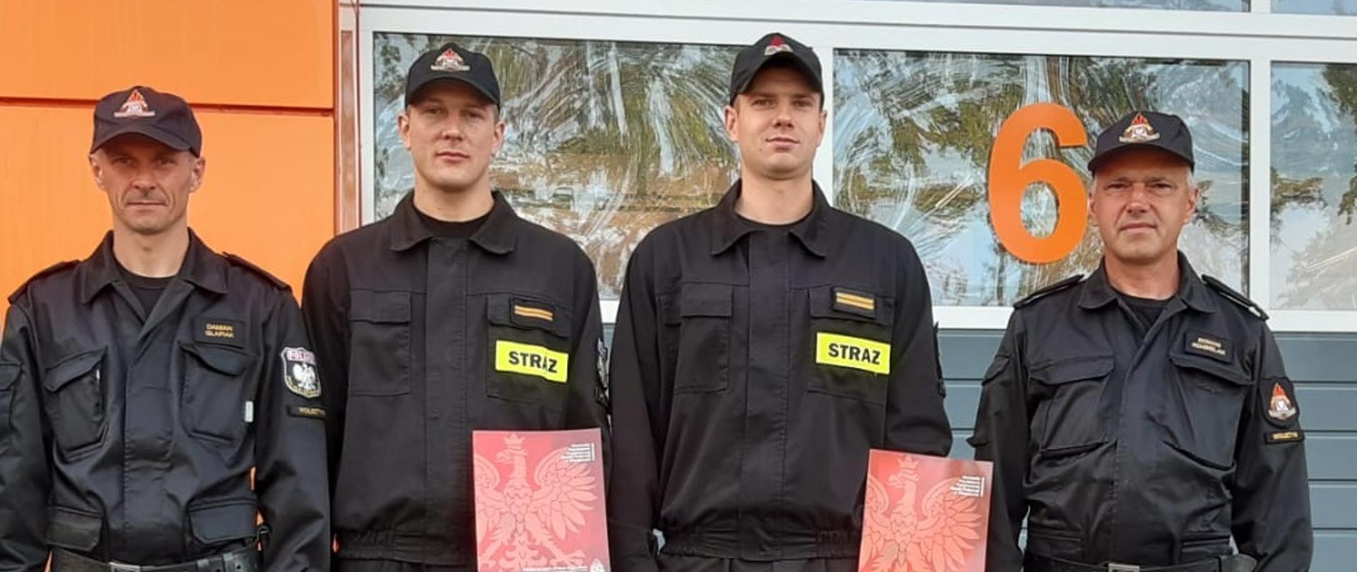 Na zdjęciu strażacy stoją na baczność i w rękach trzymają decyzję. W tle budynek KP PSP w Wolsztynie.