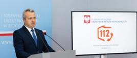 Wojewoda przemawia podczas konferencji w tle tablica z grafiką numeru 112