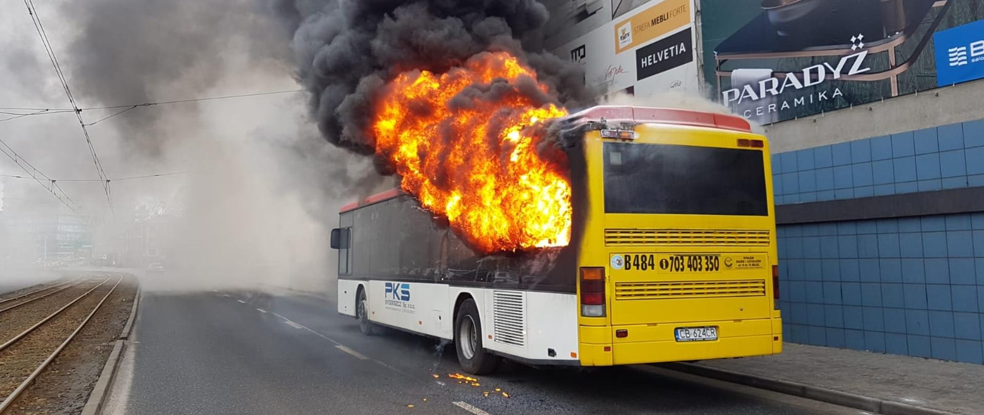 Na pierwszym planie tył płonącego autobusu, w tle budynek handlowy z zawieszonymi banerami reklamowymi. 