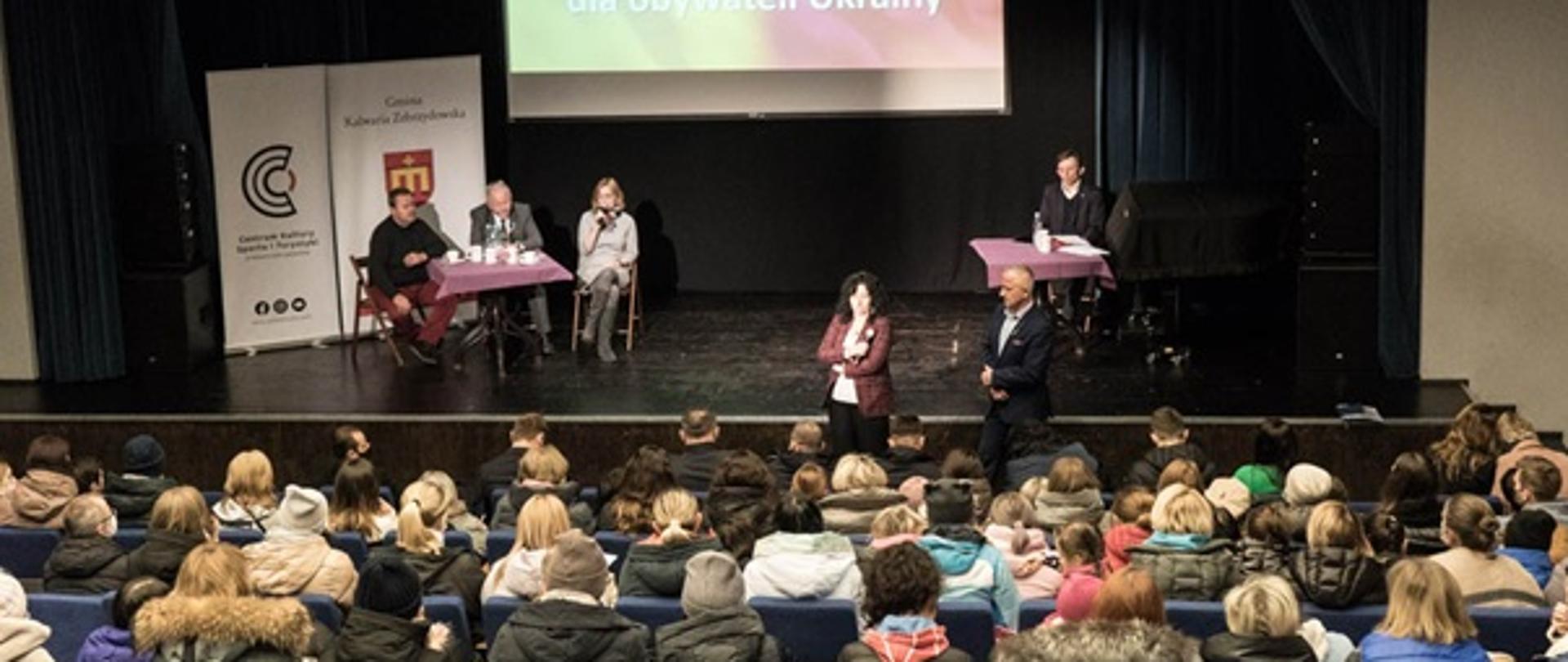 W głębi scena, na której siedzą za stolikiem trzy osoby prowadzące spotkanie. Za nimi ekran, na którym wyświetlane są aktualne informacje. Dwie kolejne osoby stoją twarzą w kierunku widowni, na której siedzą obywatele Ukrainy. 