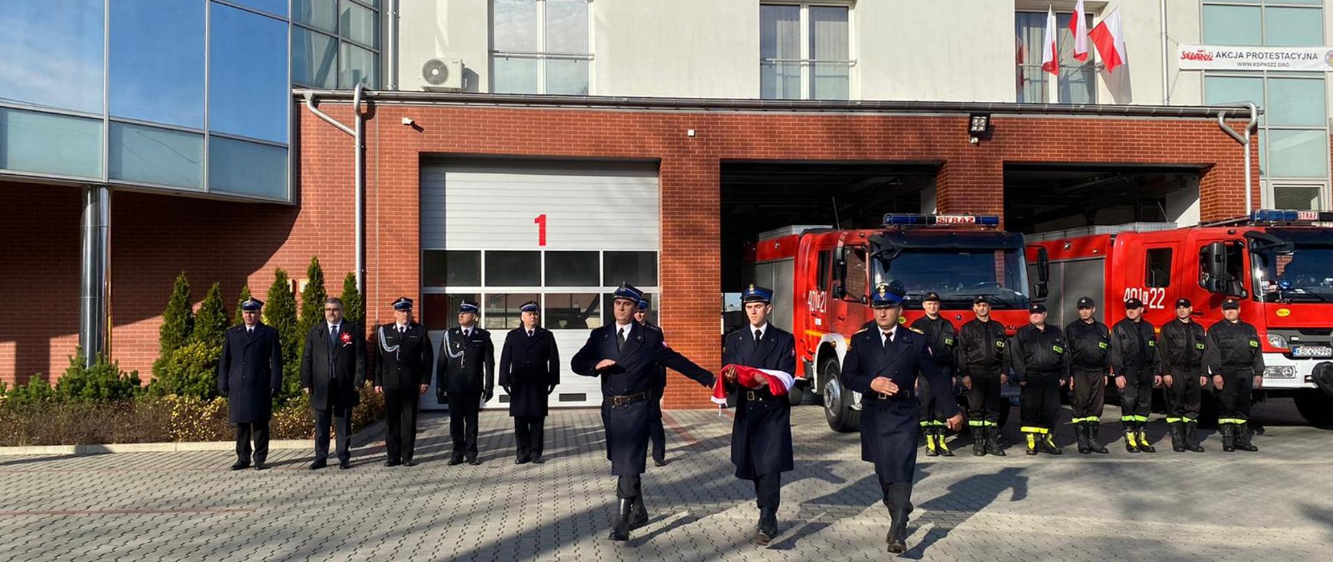 Zdjęcie przedstawia strażaków z Państwowej i Ochotniczej Straży Pożarnej w mundurach wyjściowych, strażaków ubranych ubrania koszarowe, koloru czarnego. OSP. W tle znajdują się samochody pożarnicze koloru czerwonego, bramy garażowe i budynek strażnicy