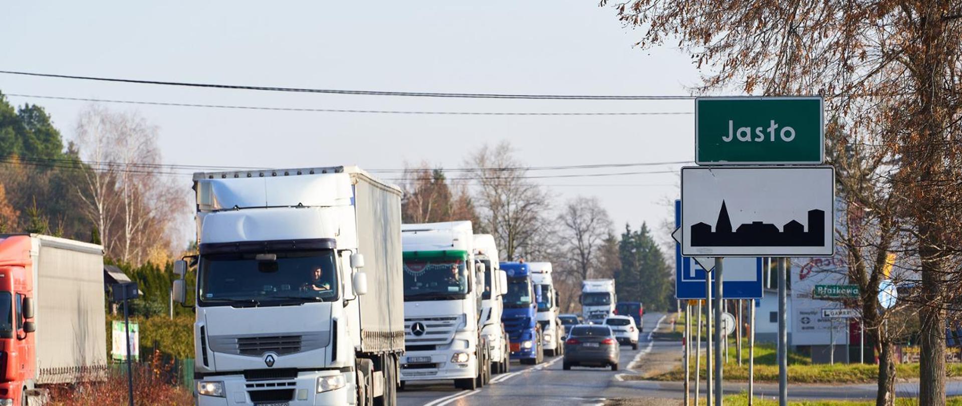 Aura jesienna. Widok na drogę, na której widać duży ruch pojazdów. Prawym pasem jadą auta osobowe, a lewym tiry. Po prawej stronie znak z napisem Jasło. W tle inne znaki drogowe ustawione wzdłuż drogi.