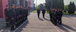 Strażacy stoją w dwóch dwuszeregach twarzami do siebie na placu apelowym. W tle budynek Komendy, drzewa i maszt flagowy.