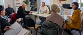 grupa osób siedzi w pomieszczeniu, na stole leżą dokumenty