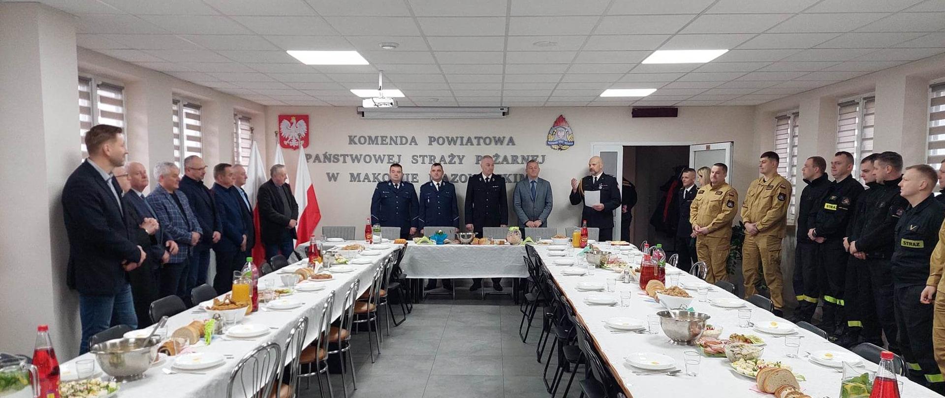 Zastępca Komendanta przemawia do funkcjonariuszy i zaproszonych gości zgromadzonych przy stole, podczas spotkania wielkanocnego, które odbywa się w świetlicy Komendy.