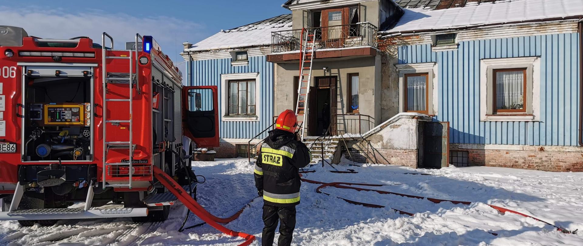 Zdjęcie przedstawia stojącego tyłem do fotografa, ubranego w strój strażacki oraz hełm, strażaka, znajdującego się na zewnątrz domu jednorodzinnego, zwróconego przodem do budynku, rozmawiającego przez krótkofalówkę. Na lewo od strażaka stoi samochód strażacki ustawiany przodem w kierunku domu. Na ziemi (na śniegu) widać rozwinięte czerwone węże strażackie wychodzące z samochodu strażackiego. Dom jednorodzinny jest w kształcie prostokąta, posiada piwnicę, parter oraz małe poddasze z balkonem od frontu domu. Budynek jest niebieski. Okna balkonowe oraz drzwi wejściowe są otwarte.