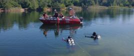 Na zdjęciu widać łódź z ratownikami, którzy podają sprzęt dwóm nurkom znajdującym się w wodzie.