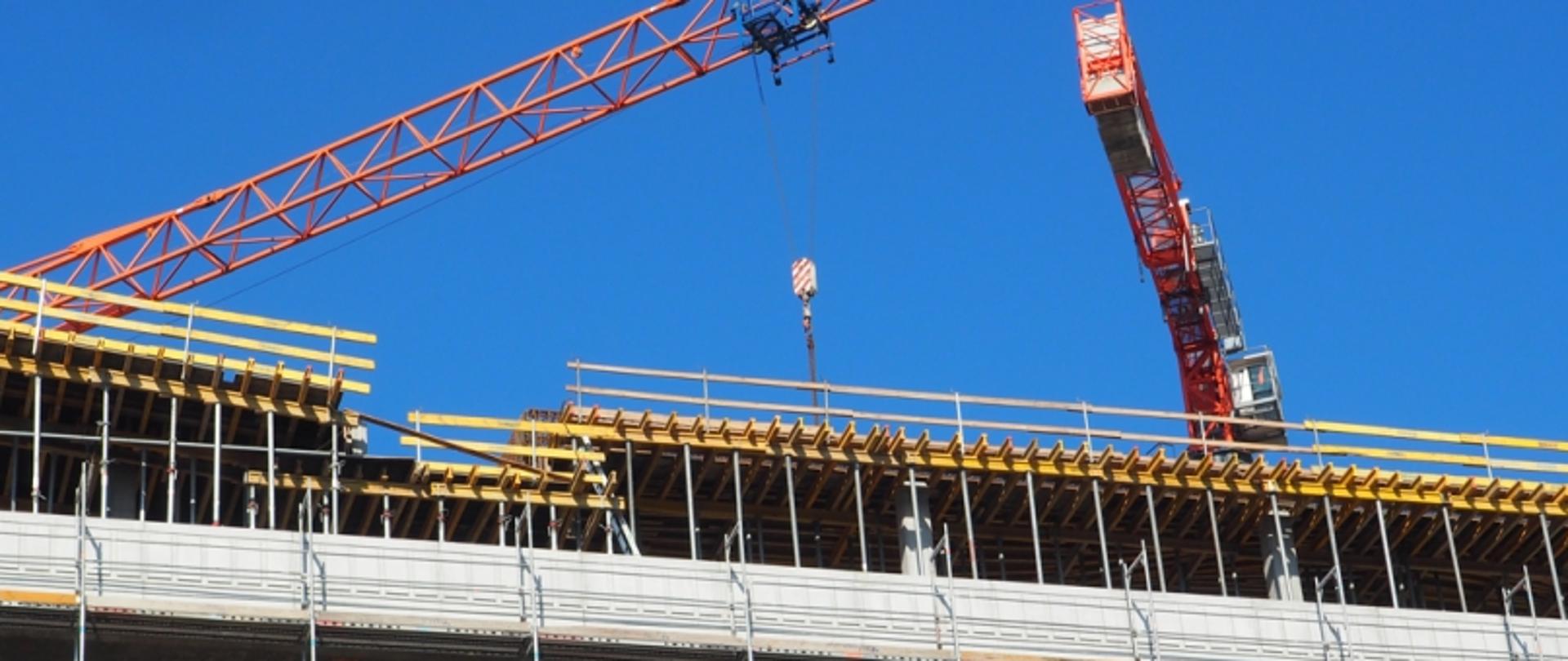 Dwa dźwigi na placu budowy przy wznoszonym budynku z rusztowaniami i belkami; niebieskie, bezchmurne niebo w tle.