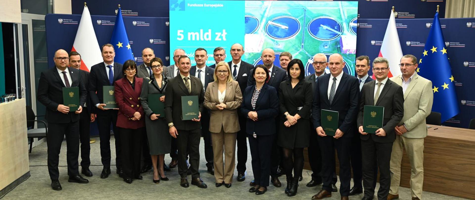 The signing of agreements by eight municipalities for European funding to support investments improving water and sewerage infrastructure. The photo shows a very large group of people, with Minister of Climate and Environment Paulina Hennig-Kloska standing at the centre. Some participants are holding folders containing the agreements. All are smiling and posing for the photo.