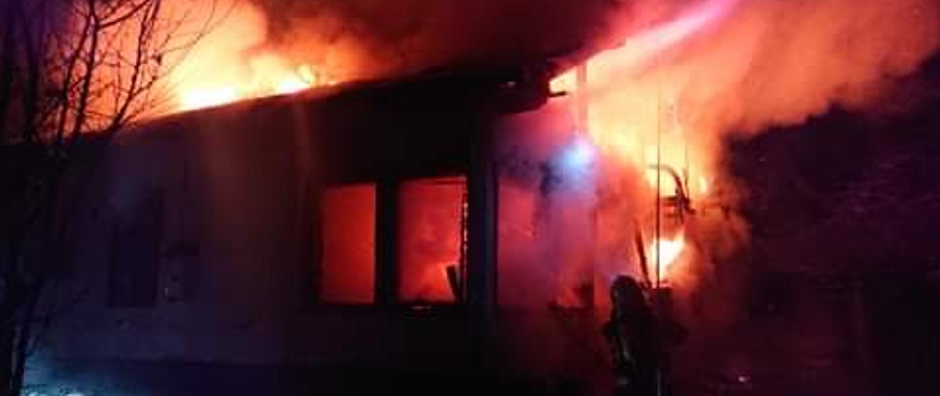 Na zdjęciu w porze nocnej widoczne samochody pożarnicze i strażacy przemieszczający się do pożaru budynku mieszkalnego. Warunki zimowe, śnieg, strażacy w trakcie rozwijania linii gaśniczych.