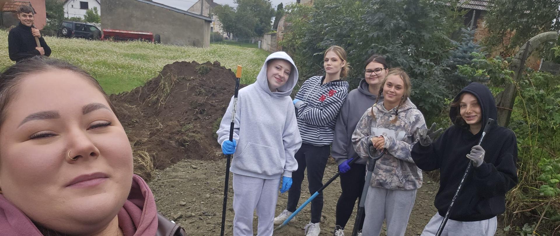 Grupowe selfie na placu budowy. Uśmiechnięci uczniowie wraz z opiekunem pozują z łopatami i grabiami na tle pryzmy ziemi i zabudowań wiejskich.