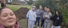 Grupowe selfie na placu budowy. Uśmiechnięci uczniowie wraz z opiekunem pozują z łopatami i grabiami na tle pryzmy ziemi i zabudowań wiejskich.