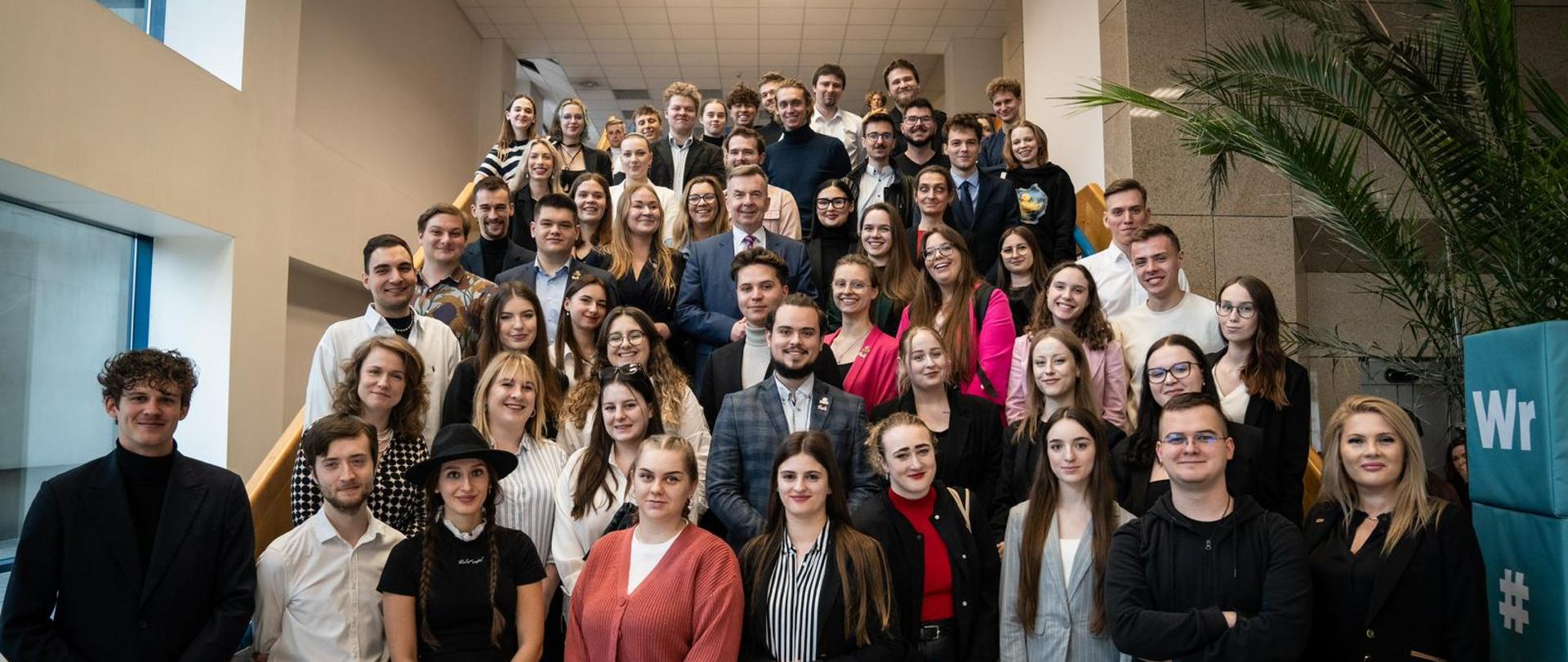 Na schodach stoi duża grupa uśmiechniętych studentów. Pośrodku grupy stoi minister Wieczorek.
