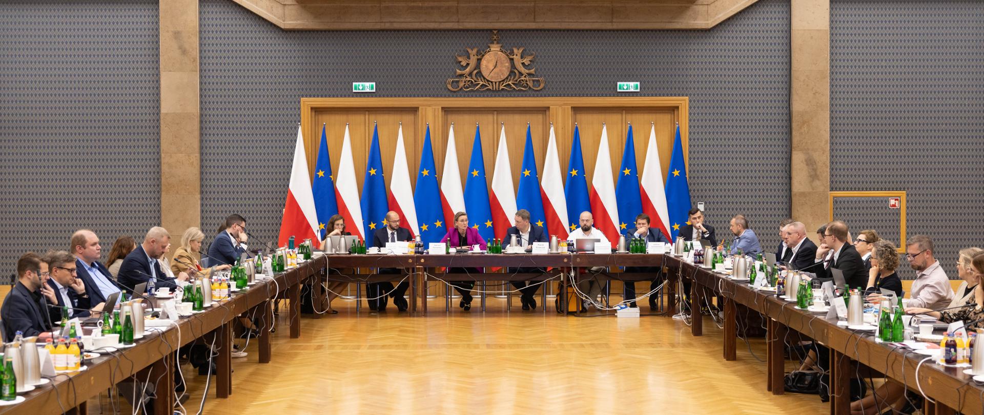 Posiedzenie Rady Działalności Pożytku Publicznego. Na zdjęciu uczestnicy wydarzenia. Szeroki kadr obejmuje widok sali i trzy strony stołu konferencyjnego, ustawionego w kształt litery "U". 