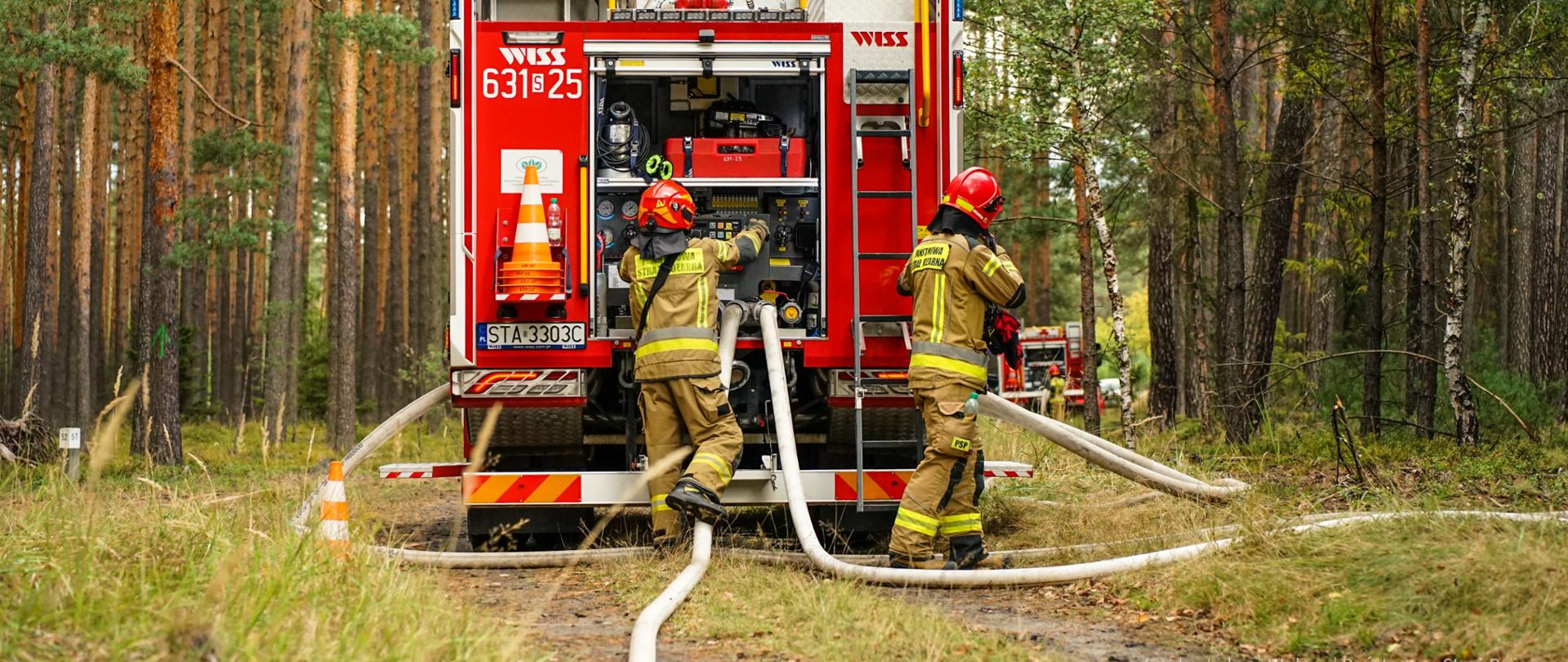 Zdjęcie przedstawia tył dużego, czerwonego pojazdu strażackiego, który stoi na polnej drodze w lesie. Dwóch strażaków, ubranych w beżowe ubrania specjalne z jaskrawożółtymi pasami odblaskowymi i w czerwonych hełmach, pracuje przy samochodzie. Jeden z nich, stojący po lewej stronie, jest zwrócony w stronę pojazdu i ma pochyloną głowę, obsługując autopompę. Drugi strażak stoi po prawej stronie wozu i trzyma w rękach szeroki, biały wąż strażacki, który jest podłączony do pojazdu. Od wozu odchodzi kilka węży strażackich. Wokół pojazdu rośnie wysoka trawa i małe drzewka. W tle widoczny jest gęsty las.