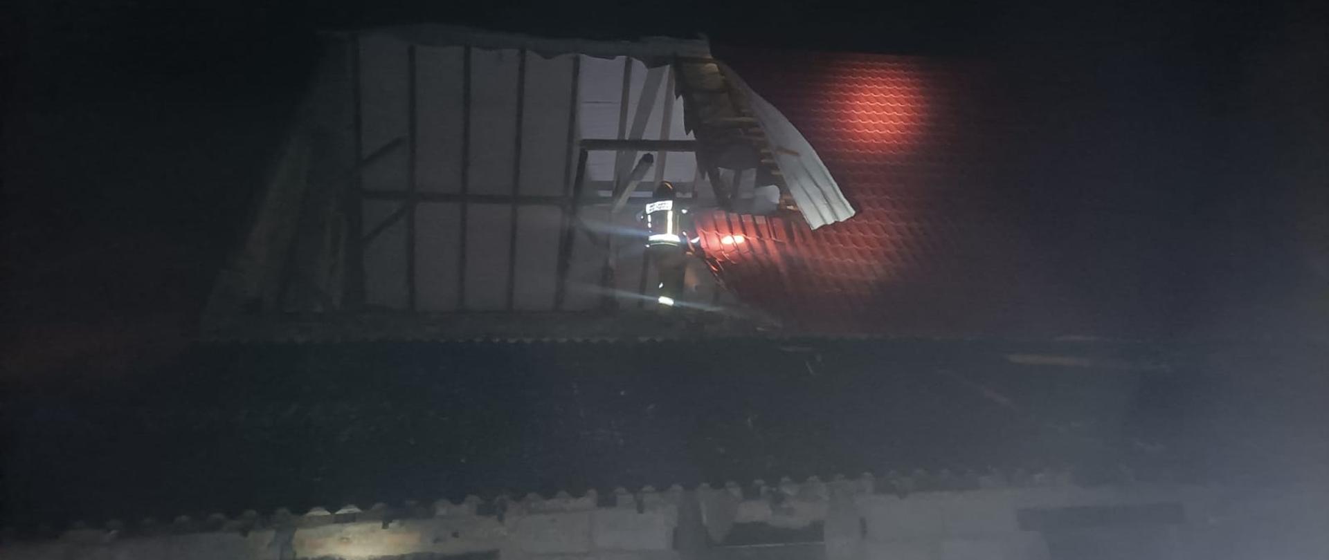 Noc. Widok na dach budynku, prawdopodobnie gospodarczego. ściany budynku nie otynkowane z budowane z bloczków w szarym kolorze. Dach pokryty blachodachówką w kolorze czerwonym. Część dachu (połowa widocznej na zdjęciu powierzchni) została zerwana. Widać wnętrze przestrzeni strychowej, w której przy krawędzi dachu stoi strażak w ubraniu specjalnym w kolorze piaskowym oraz czerwonym hełmie. Część dachu oświetlona latarkami.