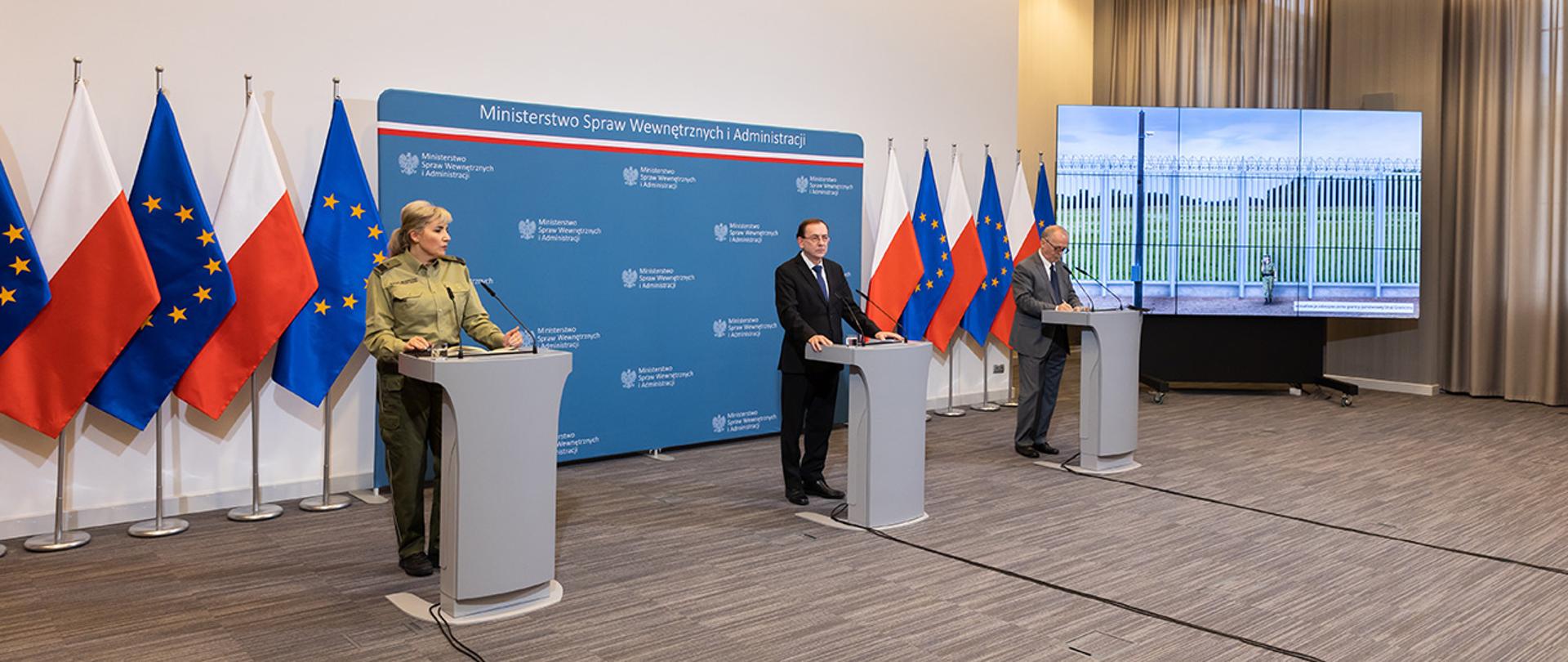 Na zdjęciu widać uczestników konferencji stojących za mównicami, widać również wyświetloną na ekranie wizualizację zapory.