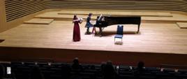 Dziewczyna w długiej ciemnoczerwonej sukience stojąc gra na altówce, lekko za nią, obok po prawej stronie kobieta gra na fortepianie.