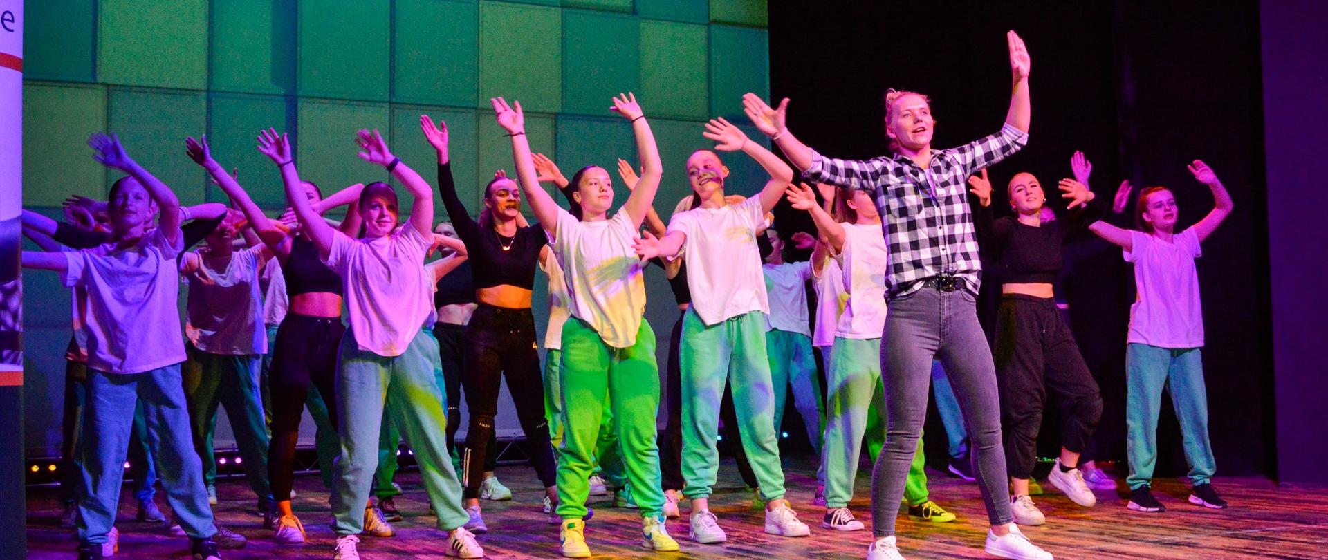 Na oświetlonej na biało i fioletowo estradzie tańczy grupa dzieci i młodzieży, z przodu kobieta w koszuli w czarno-białą kratę, wszyscy trzymają ręce w górze, za nimi ściana w zielone czworokąty.