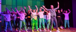 Na oświetlonej na biało i fioletowo estradzie tańczy grupa dzieci i młodzieży, z przodu kobieta w koszuli w czarno-białą kratę, wszyscy trzymają ręce w górze, za nimi ściana w zielone czworokąty.
