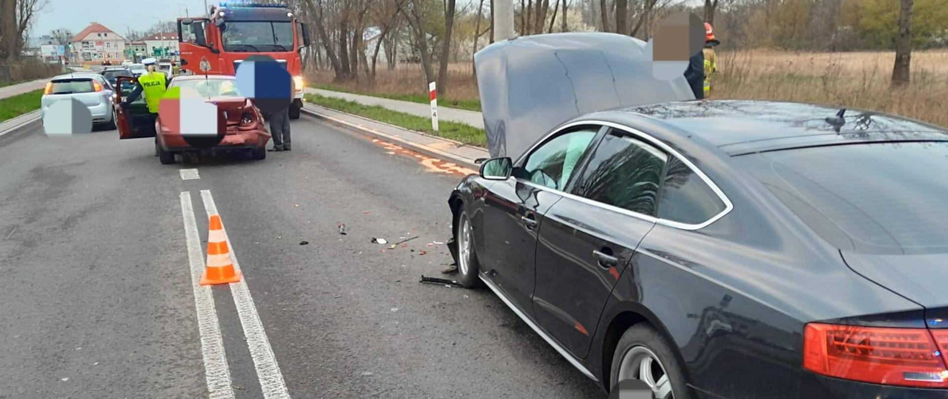 Na zdjęciu widać ciemny samochód osobowy, który ma uszkodzony przód oraz otwartą maskę. Pojazd stoi na asfaltowej drodze. Przed nim stoi bordowy samochód, który ma uszkodzony tył. W pojeździe są otwarte drzwi, przy których stoi policjantka w żółtej kamizelce i białej czapce. W tle widać czerwony samochód straży pożarnej, inne pojazdy przejeżdżające obok miejsca wypadku, budynki, drzewa i krzewy. Obok bordowego pojazdu stoi mężczyzna a obok ciemnego strażak ubrany w strój w kolorze piaskowym oraz czerwony hełm. Na środku jezdni stoi biało-pomarańczowy słupek ostrzegawczy. Jest widno.