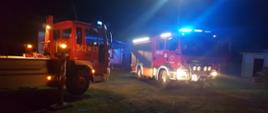 Zdjęcie wykonane w porze nocnej. Widać pojazdy straży pożarnej w kolorze czerwonym, z oznaczeniami. Na drugim planie zabudowania. Podłoże trawiaste. Występują źródła światła sztucznego z pojazdów straży pożarnej.