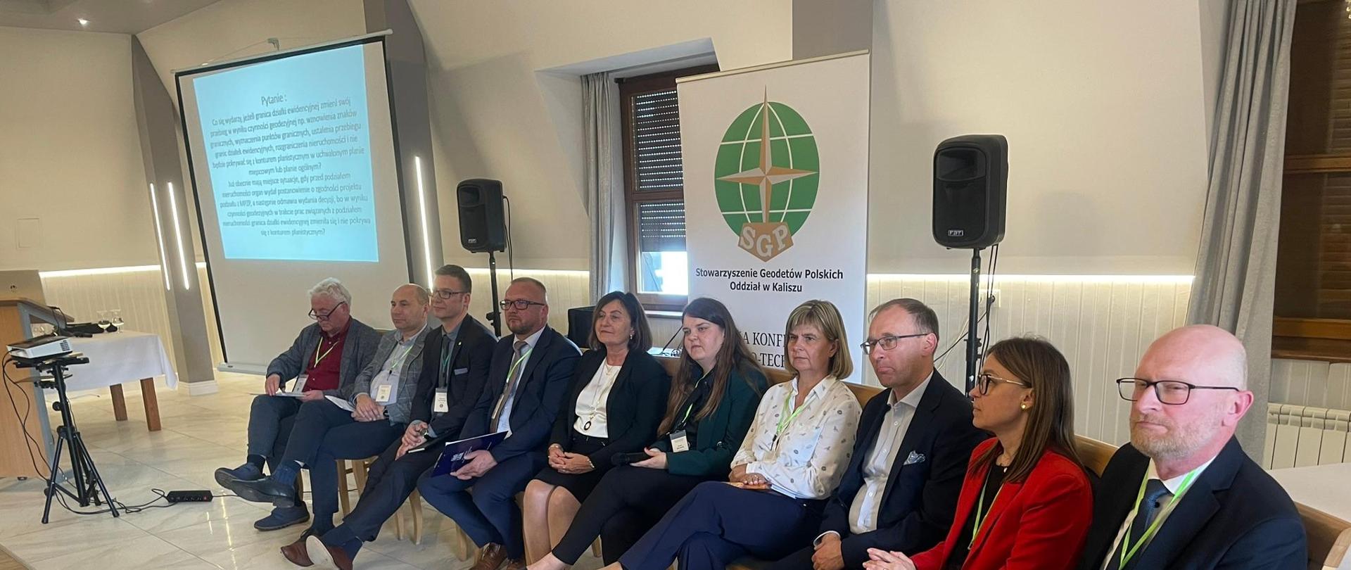 Po prawej stronie znajduje się projektor. W centralnej oraz lewej części na krzesłach siedzą Prelegenci Konferencji, w tym Pani Anna Michalik, Zastępca Dyrektora Departamentu Planowania Przestrzennego.