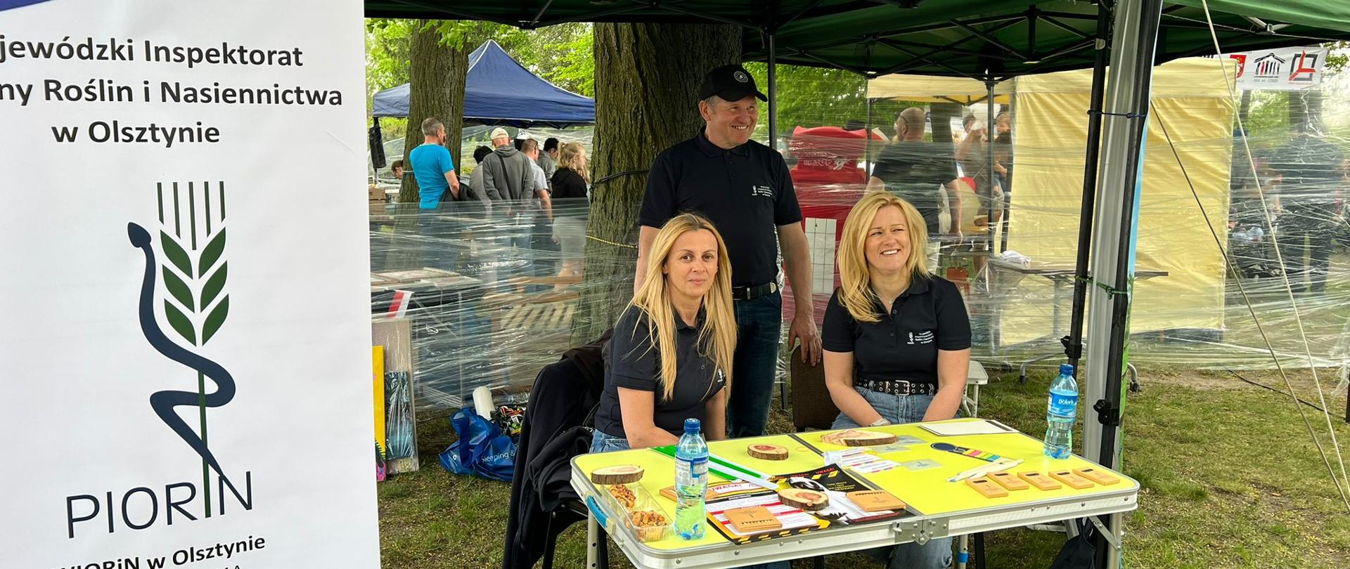 Z lewej strony rollup z napisem PIORiN, adresem i logo, pośrodku przy stoliku siedzą dwie kobiety, za nimi stoi mężczyzna, wszyscy ubrani w granatowe koszulki z logo i napisem WIORiN w Olsztynie