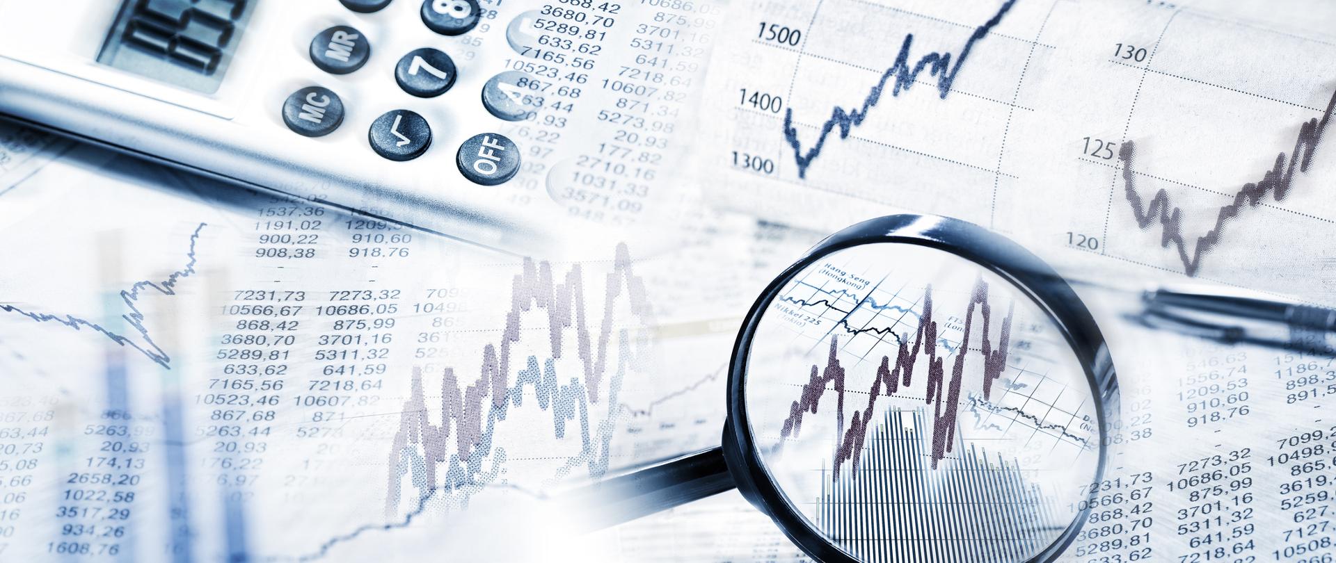 
Na obrazie lupa, wykresy giełdowe oraz kalkulator i długopis