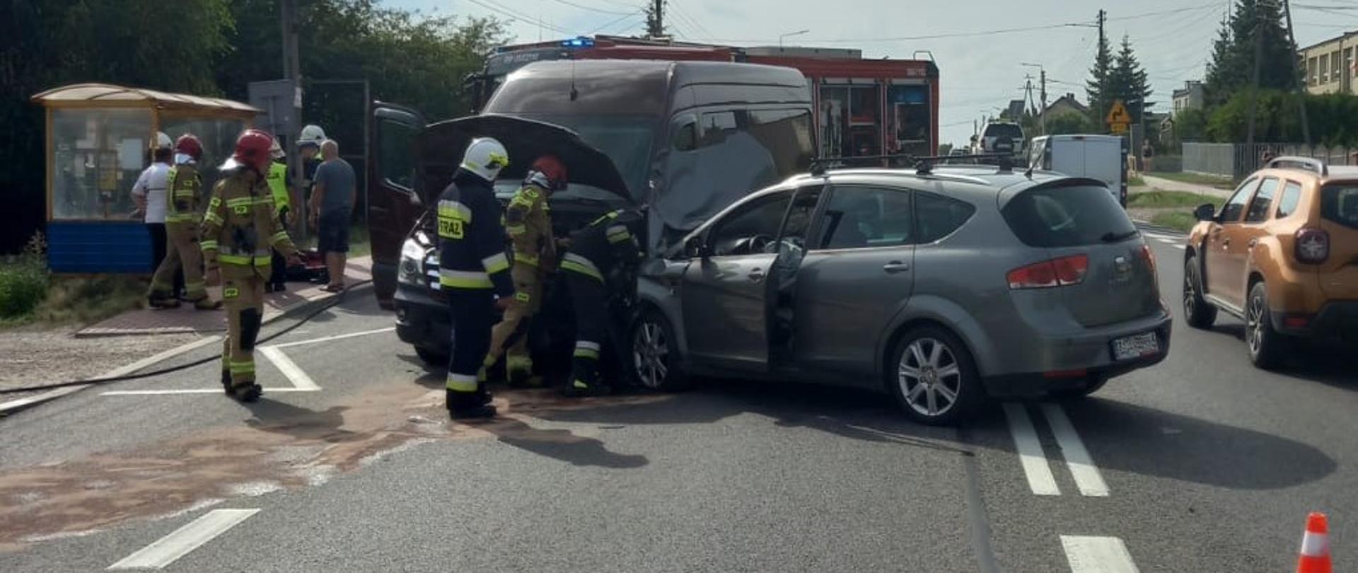 Zdjęcie przedstawia dwa auta, które uległy zderzeniu, przed nimi pracuje 3 ratowników, ruch odbywa się jednym pasem drogi. Za uszkodzonymi pojazdami widać fragment wozu strażackiego.
