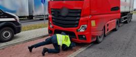 Na pierwszym planie: umundurowany inspektor opolskiej Inspekcji Transportu Drogowego pochyla się przed ciągnikiem siodłowym i sprawdza, czy z silnika nie wyciekają płyny eksploatacyjne. W tle: obok zatrzymanego zestawu w punkcie kontrolnym przejeżdża drogą samochód ciężarowy z przyczepą.