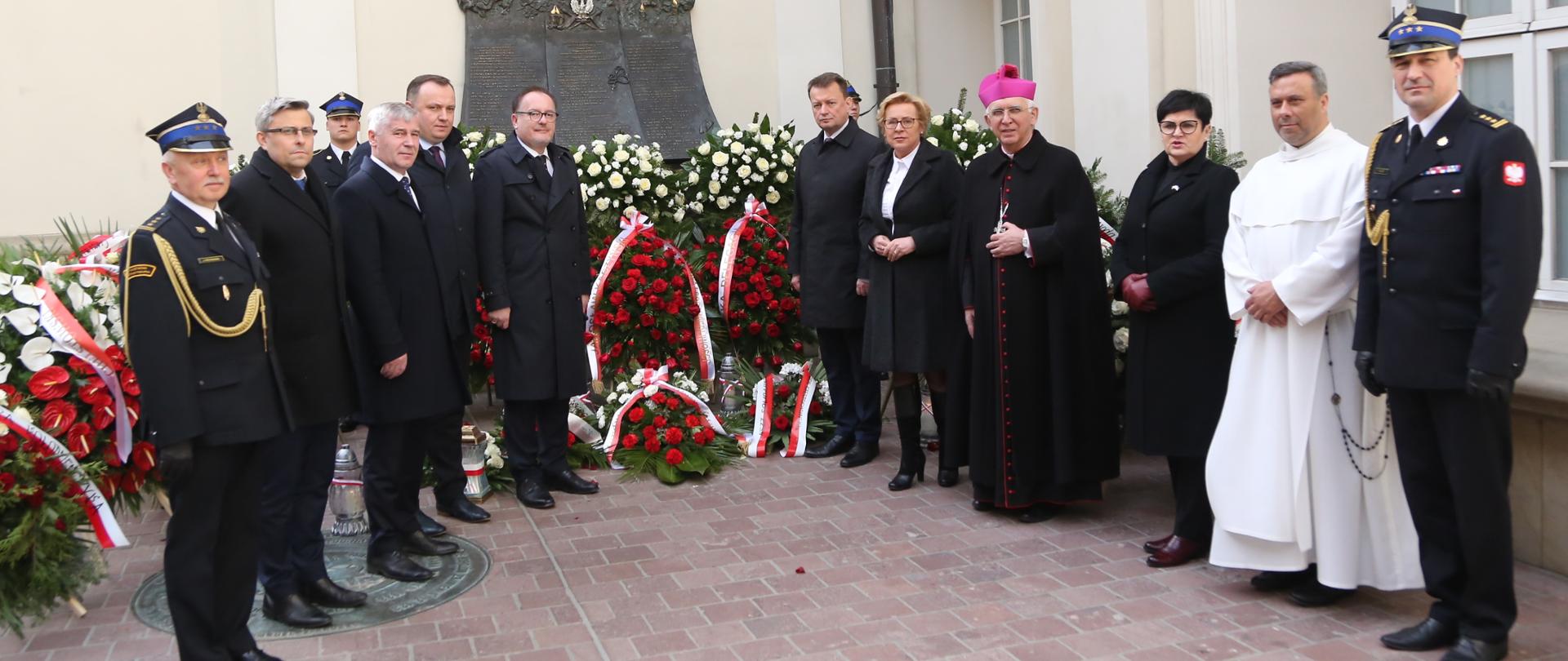 Grupowe zdjęcie uczestników uroczystości na tle pomnika upamiętniającego ofiary katastrofy, w tle złożone pod pomnikiem wieńce