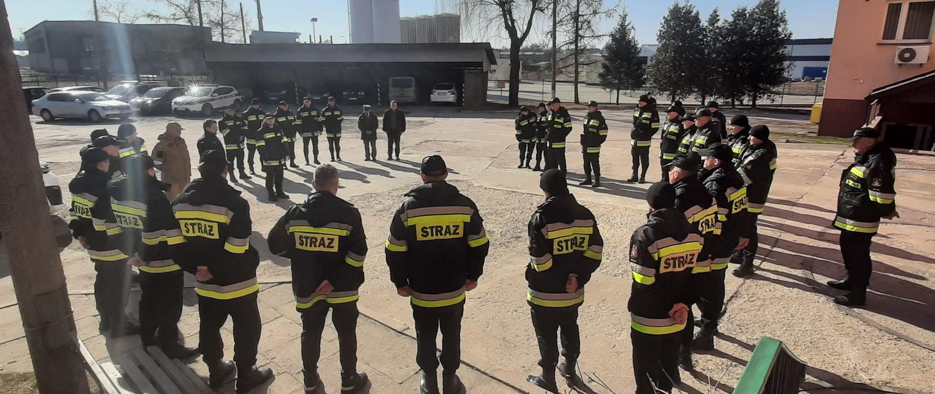 Na betonowym placu w okręgu stoi kilkudziesięciu strażaków w czarnych mundurach bojowych.