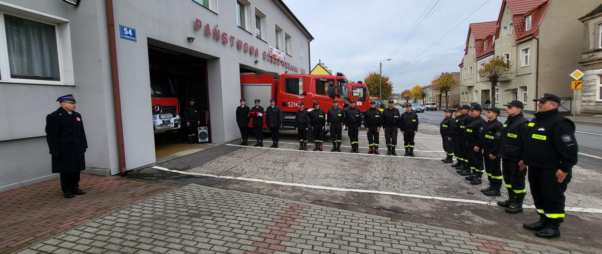 Strażacy Państwowej i Ochotniczej Straży Pożarnej w mundurach koszarowych stoją w szeregu na baczność na kształt litery L przed budynkiem Komendy Powiatowej Straży Pożarnej w Sępólnie Krajeńskim. Po lewej poczet sztandarowy oraz Komendant Powiatowy Państwowej Straży Pożarnej w Sępólnie Krajeńskim starszy brygadier Maciej Suchomski w mundurach galowych. W tle samochody ratowniczo- gaśnicze Państwowej Straży Pożarnej. 