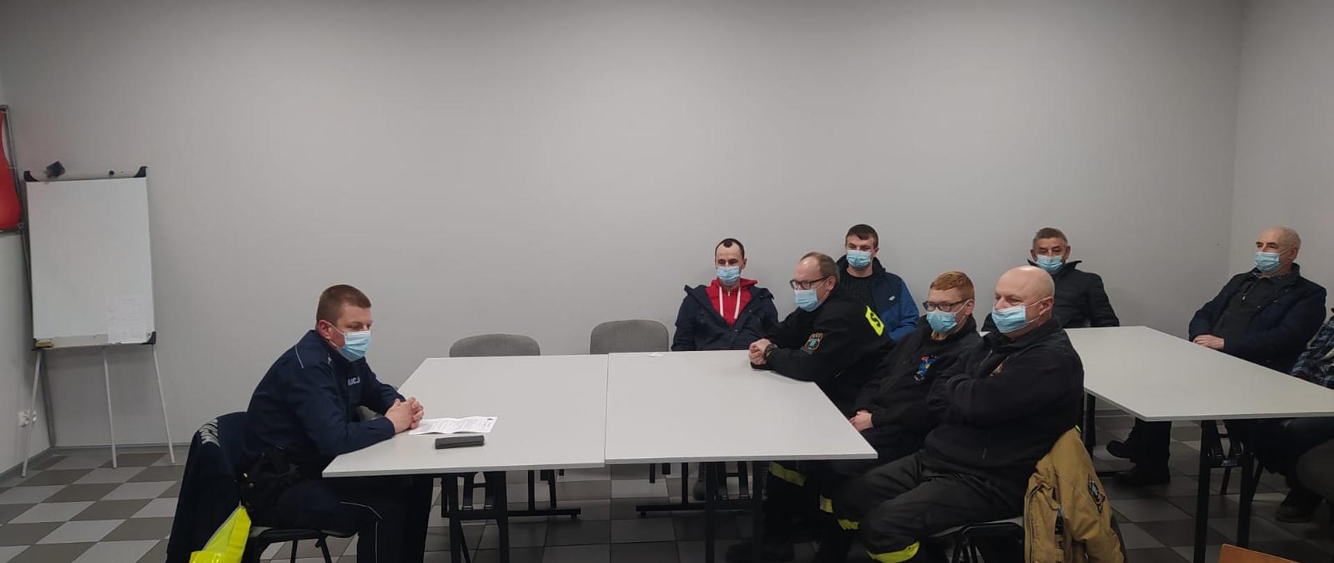 Zdjęcie przedstawia policjanta ubranego w mundur koloru granatowego siedzącego przy biurku, który prowadzi szkolenie z kierowcami OSP. i JRG.