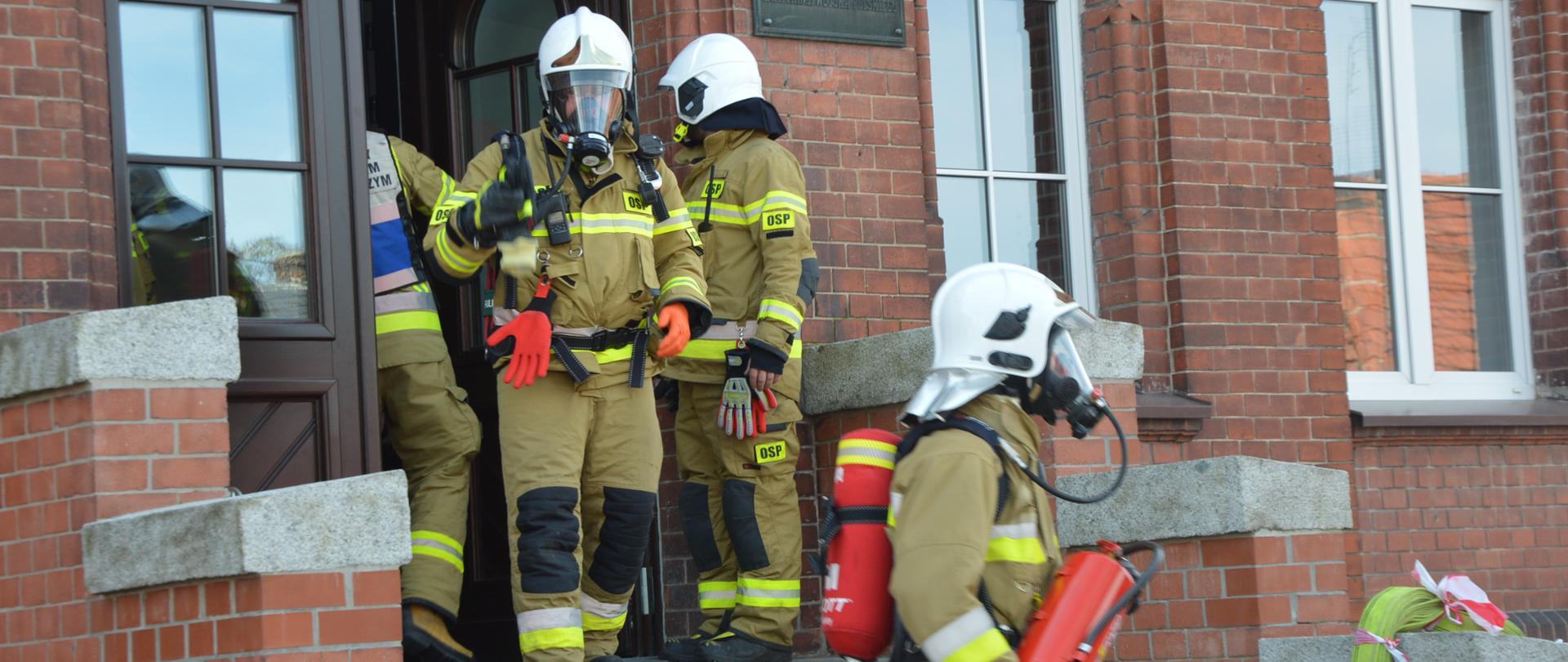 Zadymienie w przedszkolu Główne wyjście ewakuacyjnez budynku przedszkola. Czterech strażaków w ubraniach specjalnych koloru piaskowego i w białych hełmach (dwóch z nich ma założone aparaty ochrony dróg odechowych i maski) opuszcza budynek. Pierwszy z nich niesie w rękach gaśnicę. Na murku z prawej strony leży wąż tłoczny koloru żółtego.