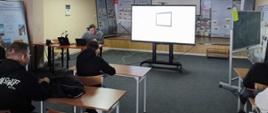 Zdjęcie przedstawia salę lekcyjną lub konferencyjną, w której odbywa się prezentacja lub lekcja.
Na pierwszym planie widoczni są od tyłu dwaj uczestnicy siedzący przy drewnianych biurkach. Jedna z osób ubrana jest w czarną bluzę z jasnym napisem na rękawie.
W głębi sali, z lewej strony, siedzi prelegent przy biurku z otwartym laptopem.
Centralnym elementem tła jest duży, płaski monitor stojący na środku pomieszczenia, na którym wyświetlony jest prosty, biały prostokąt.
Na ścianach wiszą plakaty informacyjne. Na jednym z nich, nad prelegentem, znajduje się napis ""ZESPÓŁ SZKÓŁ CENT..."", a dalej ""W KOWALU"".
Oświetlenie sali jest jasne, pochodzące z lamp sufitowych. Na podłodze leży szary dywan.