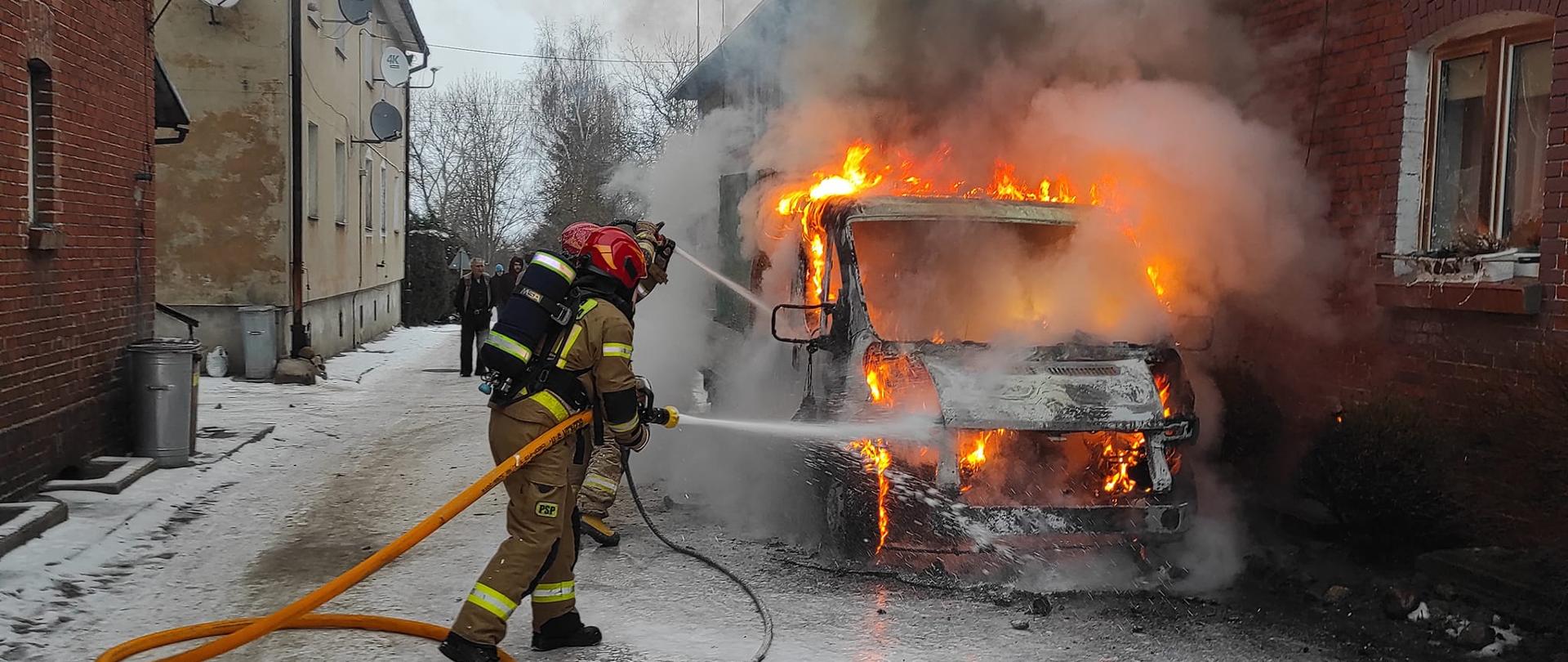 Przy budynku z czerwonej cegły pali się samochód osobowy, duże płomienie. Strażacy z wężami, gaszą pożar. Unosi się dużo dymu.