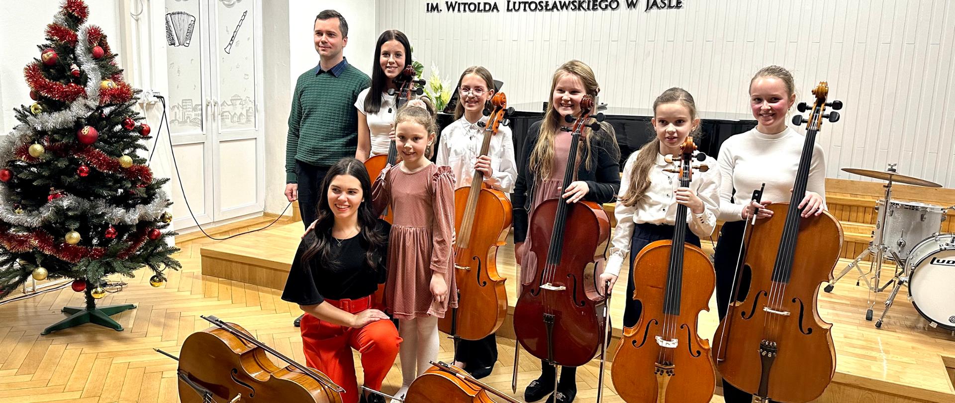 Zdjęcie przedstawia 8 osób, w tym 6 uczennic klasy wiolonczeli oraz dwójkę nauczycieli - wiolonczelistkę i pianistę. Uczennice trzymają swoje instrumenty. Wszyscy stoją blisko siebie i są uśmiechnięci. W tle widać logo Państwowej Szkoły Muzycznej I stopnia imienia Witolda Lutosławskiego w Jaśle. Po lewej stronie uczestników koncertu stoi kolorowa choinka. 