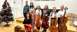 Zdjęcie przedstawia 8 osób, w tym 6 uczennic klasy wiolonczeli oraz dwójkę nauczycieli - wiolonczelistkę i pianistę. Uczennice trzymają swoje instrumenty. Wszyscy stoją blisko siebie i są uśmiechnięci. W tle widać logo Państwowej Szkoły Muzycznej I stopnia imienia Witolda Lutosławskiego w Jaśle. Po lewej stronie uczestników koncertu stoi kolorowa choinka. 