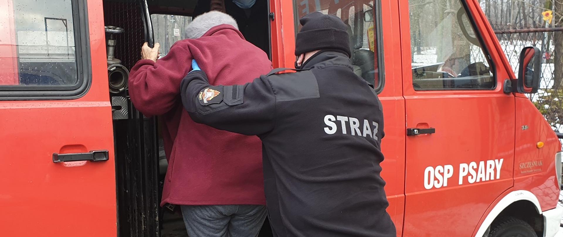 Zdjęcie przedstawia druha OSP w ubraniu koszarowym pomagającego wsiąść starszej osobie do samochodu pożarniczego. Zdjęcie wykonano podczas transportu na szczepienie.